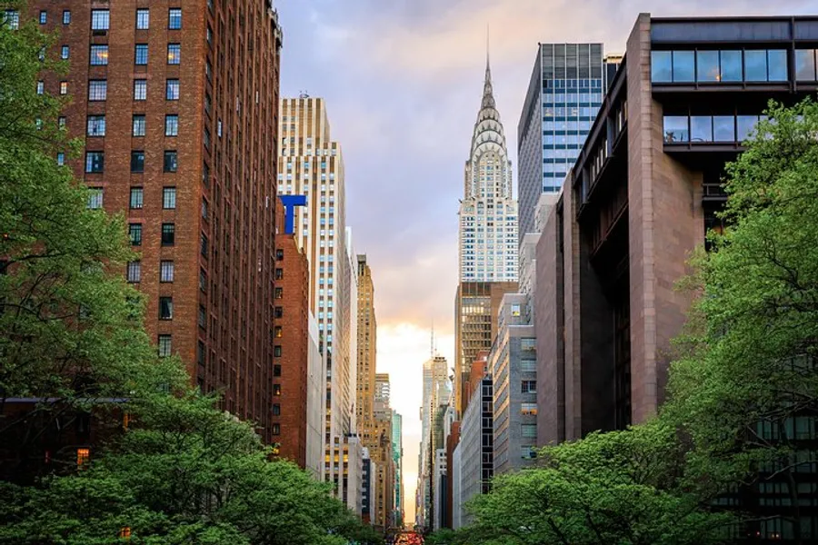 The image depicts a lush, tree-lined street in a bustling city with the iconic Chrysler Building standing tall amidst surrounding skyscrapers against a backdrop of a dramatic evening sky.