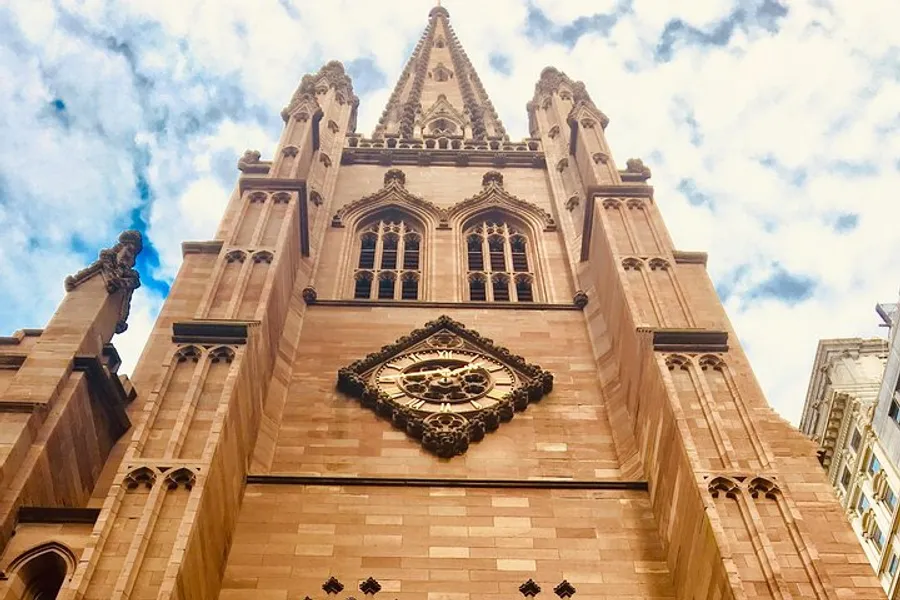 The image shows the upward view of a grand, ornate church facade with a large circular clock, set against a partly cloudy sky.