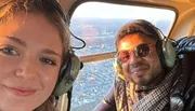 Two people are wearing headphones and smiling for a selfie inside a helicopter with a cityscape visible through the window in the golden light of sunset.