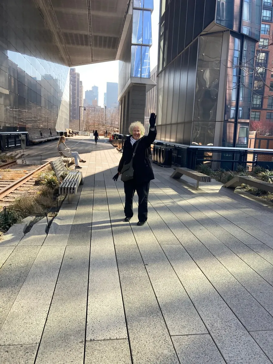 A person with light hair waves cheerfully while walking along an urban highline park, with high-rise buildings and seated individuals in the background.