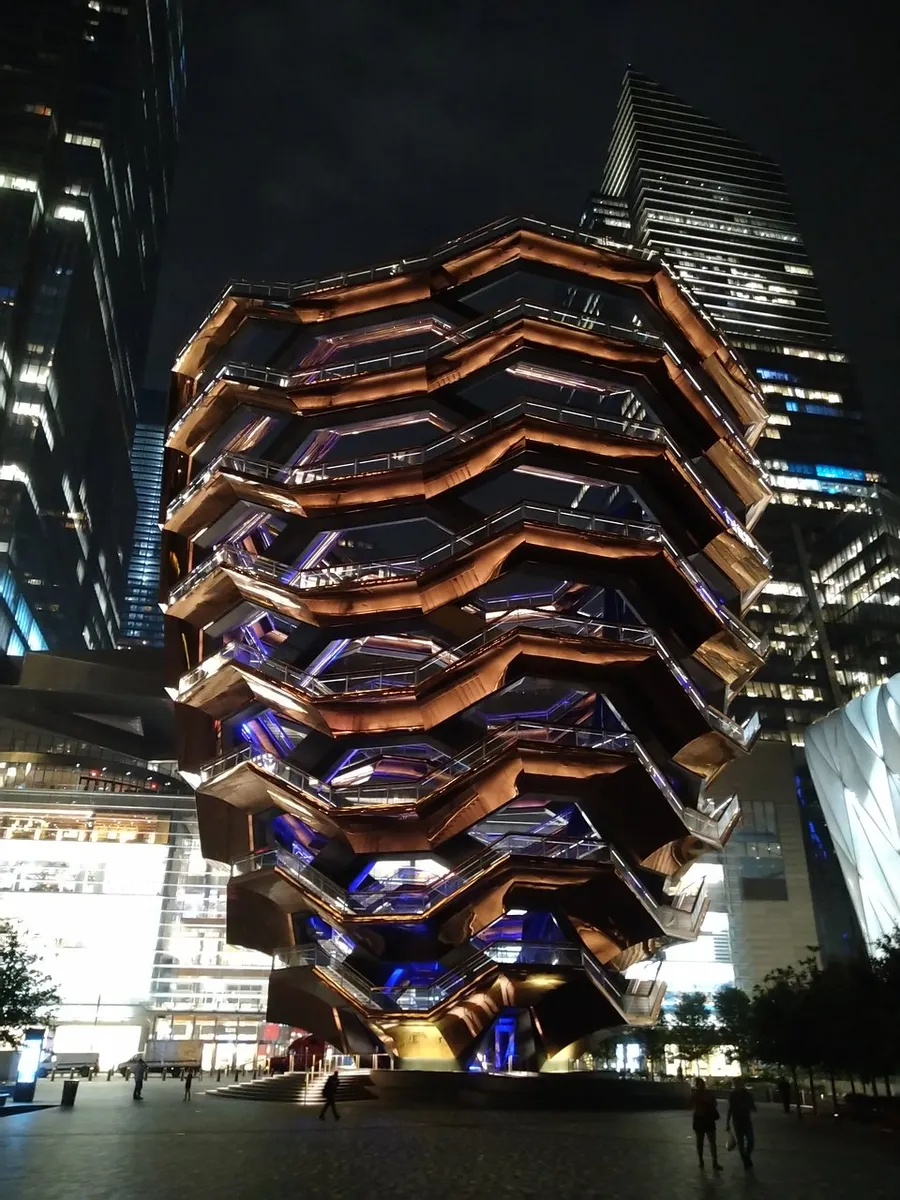 The image showcases a striking, honeycomb-like structure illuminated at night with modern skyscrapers in the background, highlighting a blend of innovative architecture and urban nightlife.