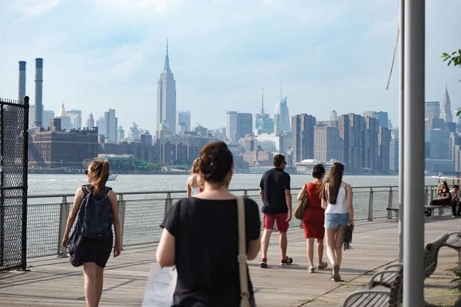 People are walking along a riverside boardwalk with a view of the New York City skyline in the background.