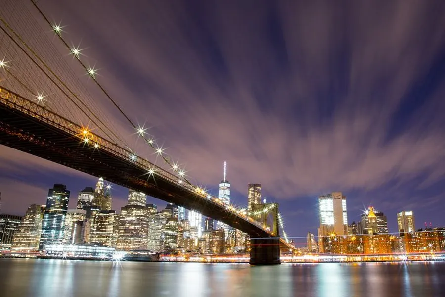 The image shows a long exposure night shot of the Brooklyn Bridge with radiant stars of light and streaky clouds above, set against the backdrop of the illuminated New York City skyline.