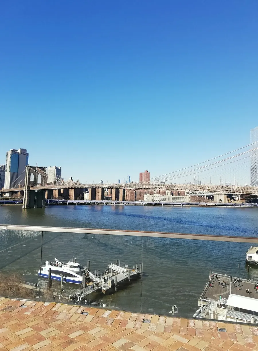 This image captures a sunny day view of a bridge over a river with docked boats and a city skyline in the background.