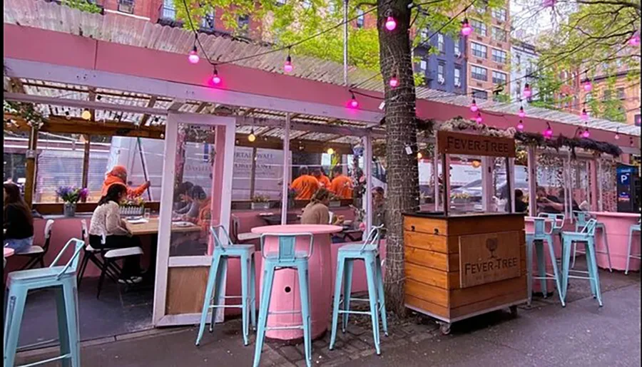 The image shows a vibrant outdoor seating area of a restaurant with pink and turquoise decor and patrons dining under a canopy adorned with hanging lights.