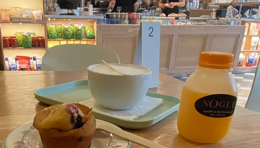 A tray with a large cup of cappuccino, a muffin, and a bottle of orange juice sits on a table in a cafe with a counter and products in the background.