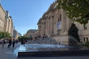 The image shows a bustling scene outside the Metropolitan Museum of Art with visitors and a fountain in the foreground on a bright sunny day.
