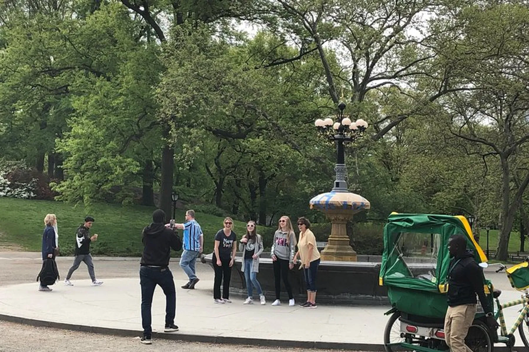 A person is taking a photo of a group next to an ornate lamp post in a park with a pedicab in the foreground and lush greenery in the background.