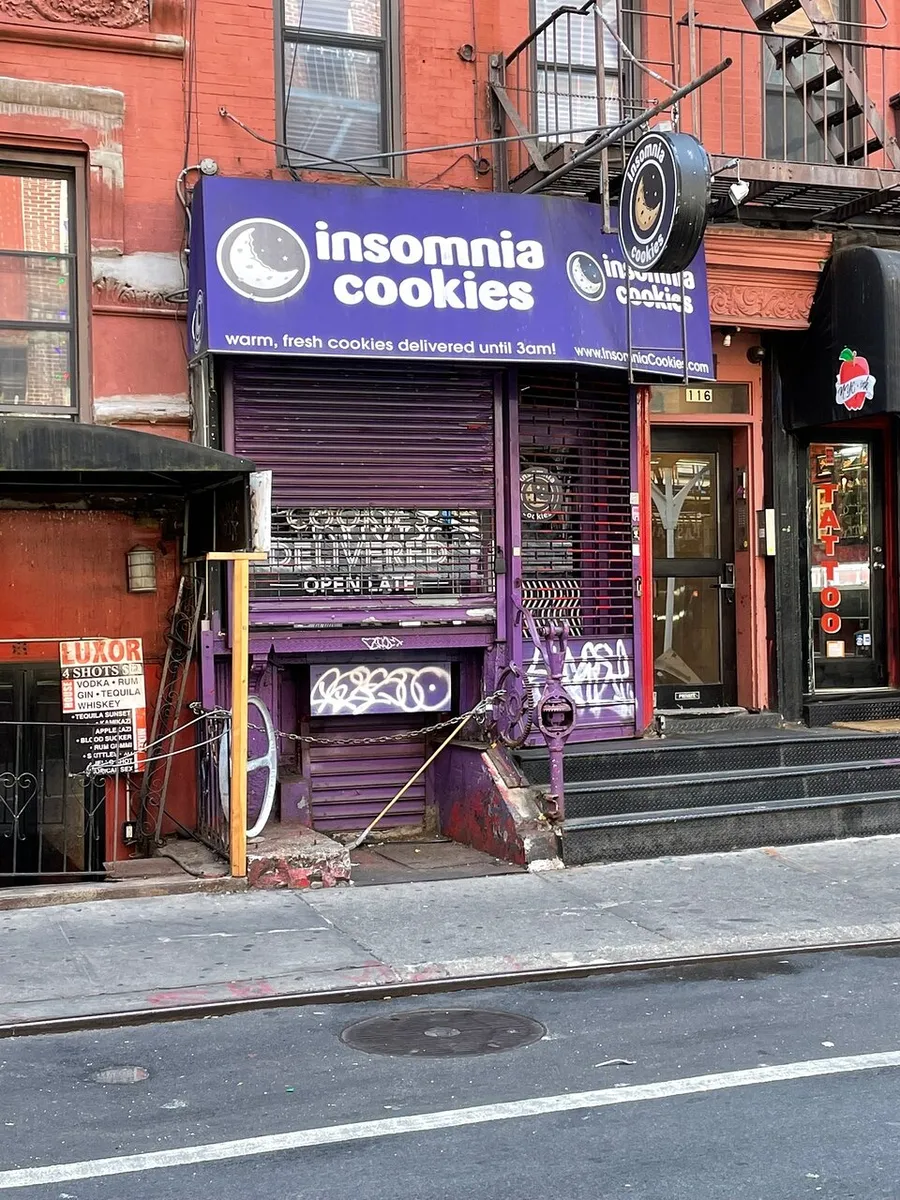 The image shows the storefront of Insomnia Cookies with its distinctive purple awning and signage, advertising warm, fresh cookies delivered until 3 am.