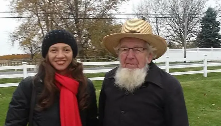 The image shows an elderly man with a straw hat and a long white beard standing next to a smiling younger woman wearing a black beanie and a red scarf, with a white fence and trees in the background.