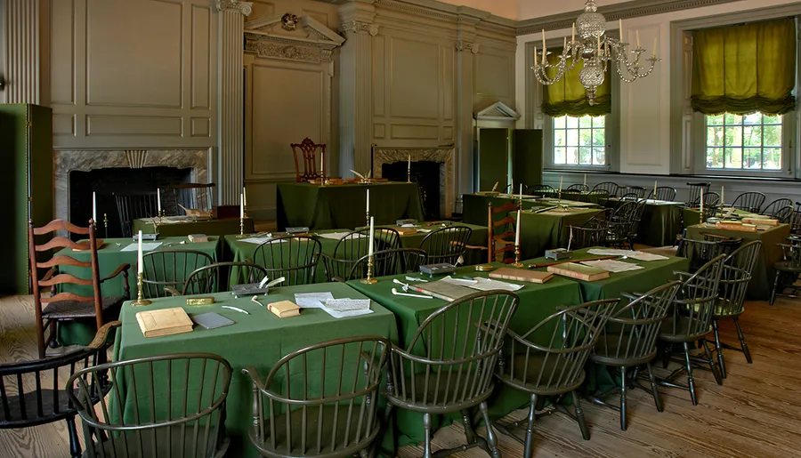 The image shows a historical room with green tablecloths, wooden chairs, and documents, suggesting it is a carefully recreated setting from a significant past event.