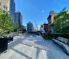 The image displays a sunny urban park scene with modern and historical architecture lining an elevated walkway peppered with greenery and benches leading towards a city skyline