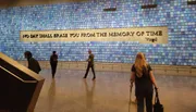 The image shows people walking past a wall with a mosaic and the Virgil quote 