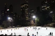 Skaters enjoy an evening on an outdoor ice rink with the illuminated backdrop of a city skyline.