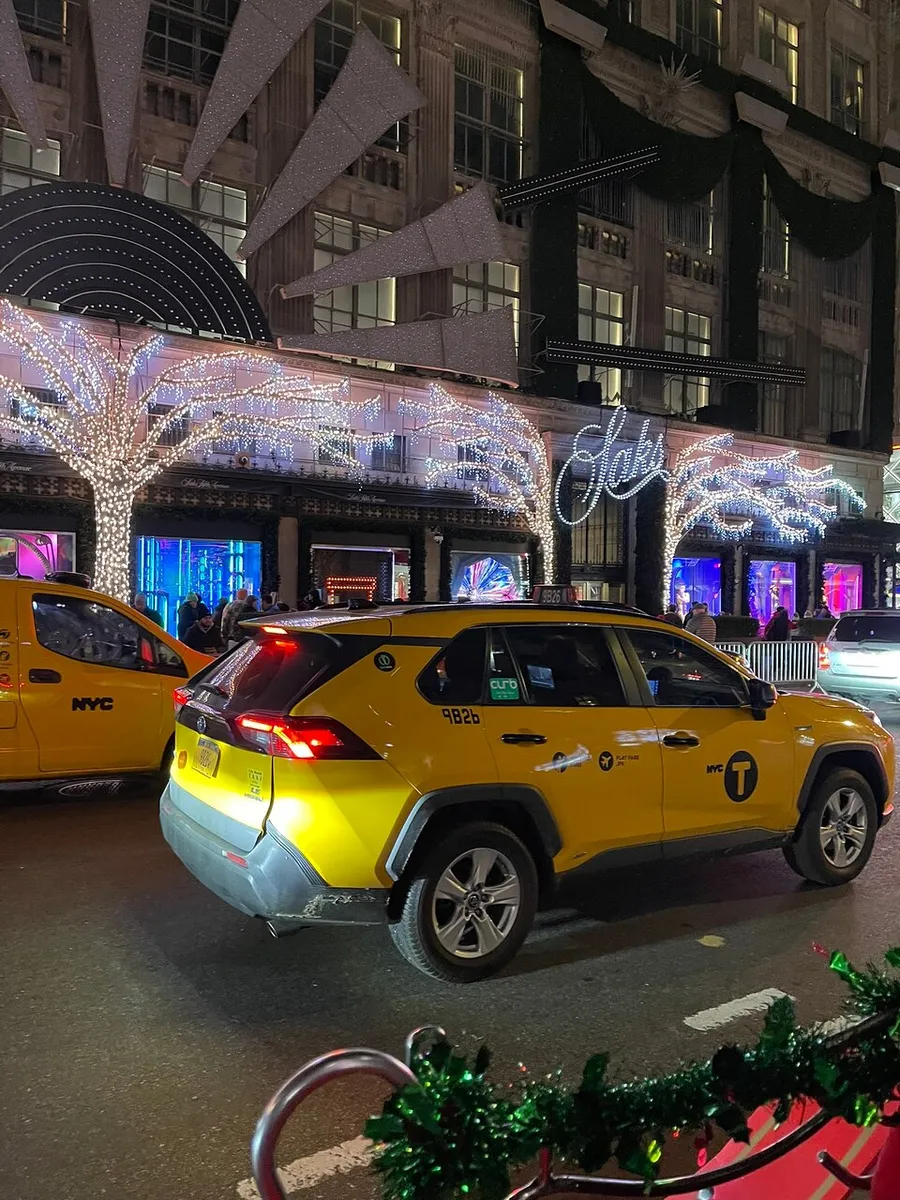 An iconic yellow NYC taxi is passing by a festively decorated building with large illuminated stars and glowing trees at night.