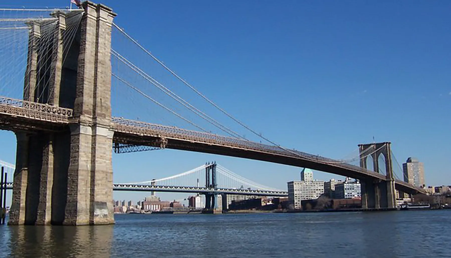 The image shows the Brooklyn Bridge, an iconic suspension bridge spanning the East River between Manhattan and Brooklyn in New York City, with the Manhattan Bridge in the background.