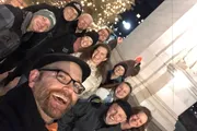 A group of smiling people is taking a cheerful group selfie at night with twinkling lights in the background.