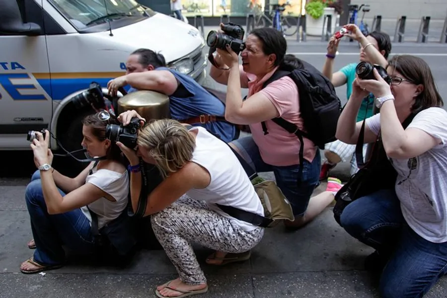 A group of photographers is crouched down, aiming their cameras towards something of interest out of the frame, near a police vehicle on the street.
