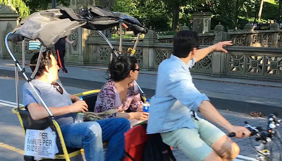 Three people are seated on a makeshift bike-powered vehicle with an umbrella on a sunny day, while the person in the front gestures towards something out of frame.