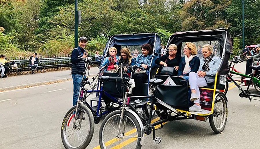 A group of people enjoys a ride in pedicabs, as the drivers stand by, in a tree-lined outdoor setting.