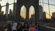 Pedestrians are enjoying a walk on the Brooklyn Bridge at sunset with the Manhattan skyline in the background.