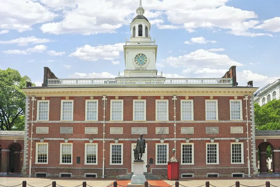 The image shows Independence Hall with a clock tower in Philadelphia, a historical building against a blue sky, with a statue standing prominently in its courtyard.
