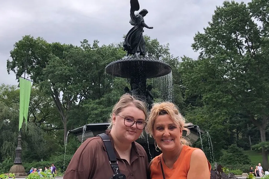 Two women are smiling for a photo in front of a fountain with a statue on it in a park setting.