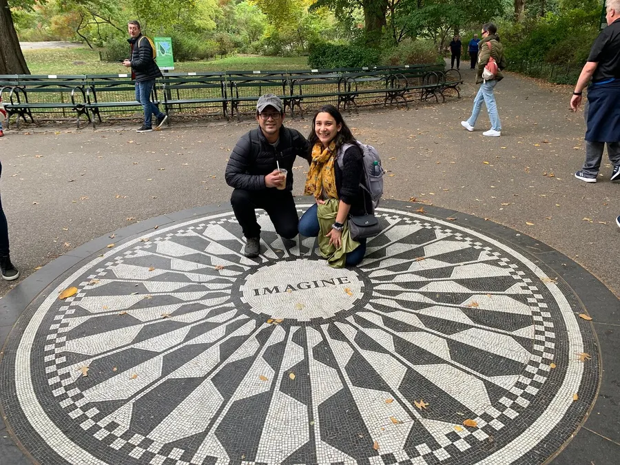 Two people are smiling and posing for a photo on the Imagine mosaic memorial in Central Park.