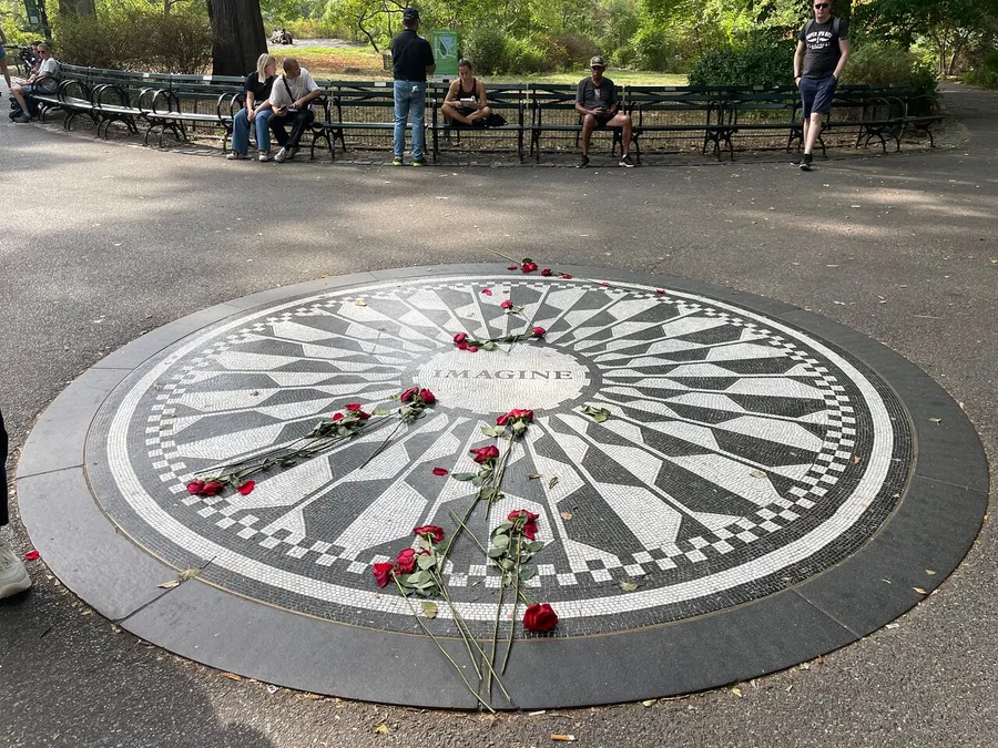 The image shows a mosaic with the word IMAGINE at its center, adorned with scattered red roses, surrounded by people sitting and standing around it in what appears to be a park.