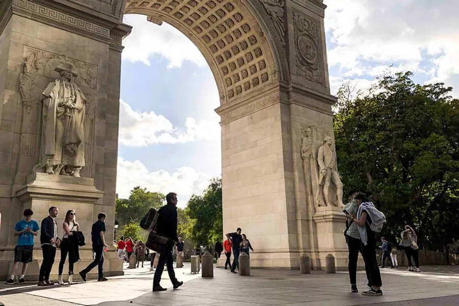 The image shows people passing by and congregating near a large, ornate archway on a sunny day, likely in a city park.