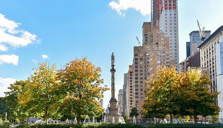 The image shows a cityscape with autumn-colored trees in the foreground, a monumental column with a statue on top in the middle, and a backdrop of mixed architecture under a blue sky with scattered clouds.