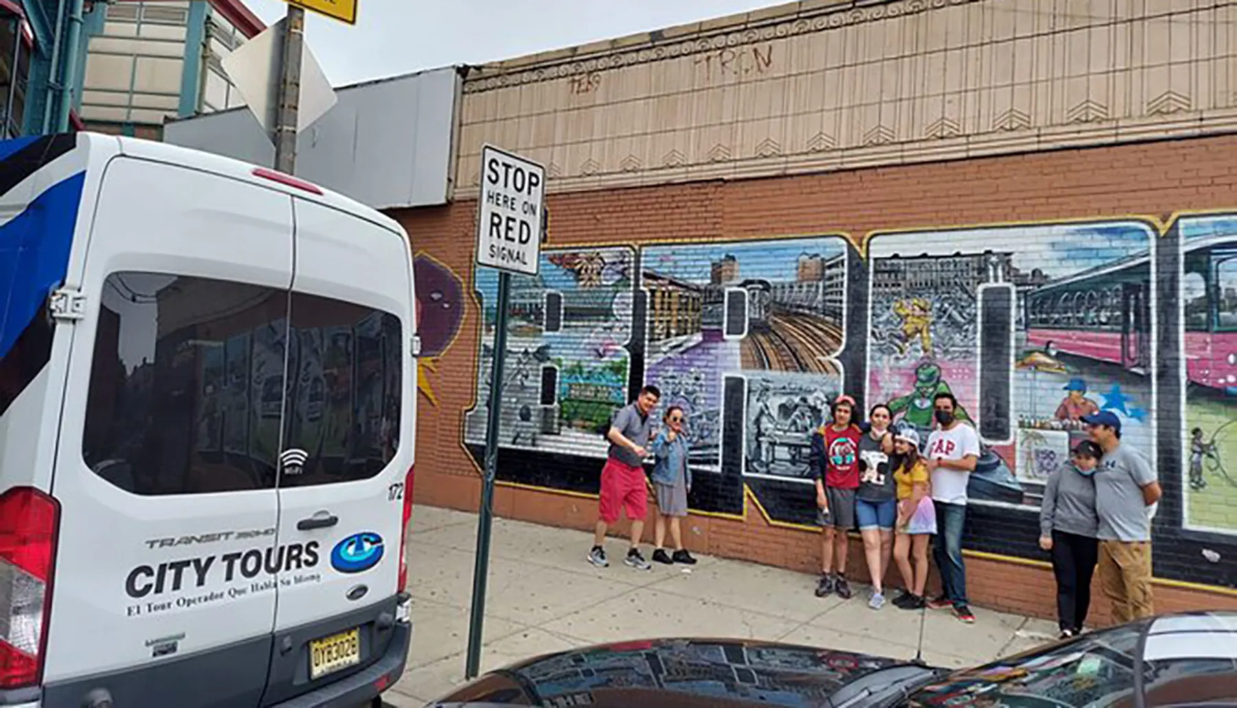 A group of people poses for a photo in front of a colorful urban mural while a city tour van is parked nearby.