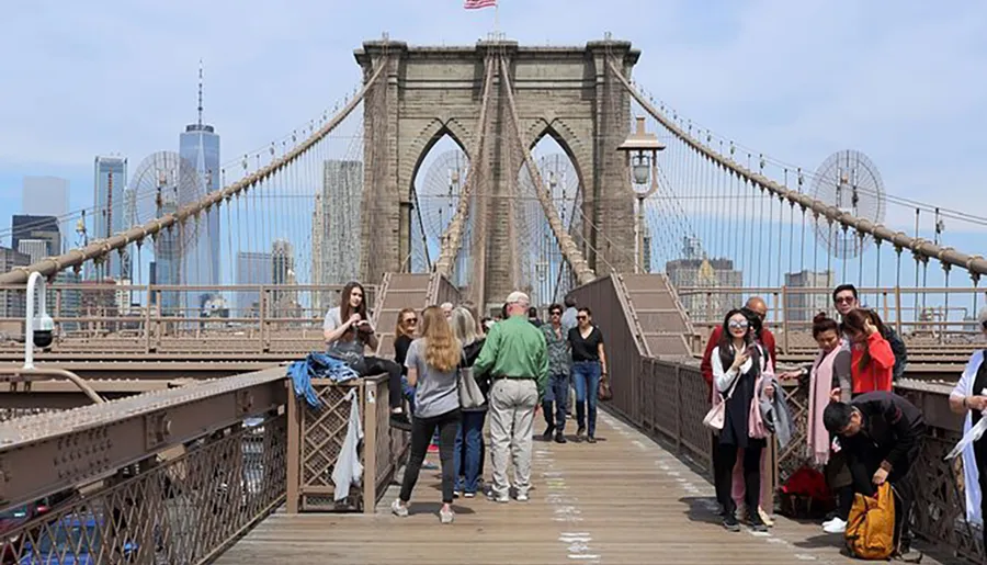 The image shows pedestrians walking and taking photos on the Brooklyn Bridge, with the New York City skyline in the background.