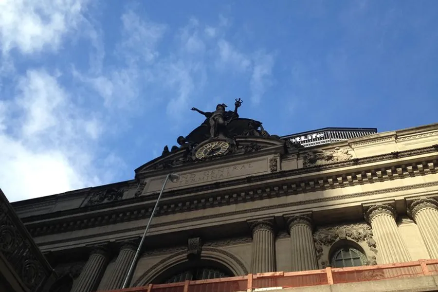 The image shows the upper facade of a grand classical building with the inscription Grand Central Terminal beneath an ornate sculpture and clock, framed against a partly cloudy sky.