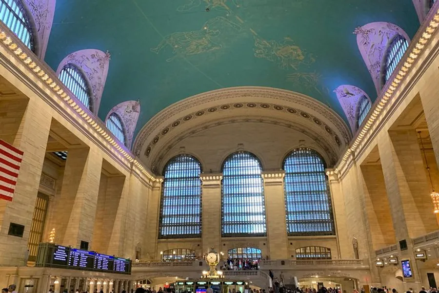 The image shows the interior of Grand Central Terminal in New York City, with its expansive main concourse, large windows, iconic celestial ceiling, and bustling atmosphere.