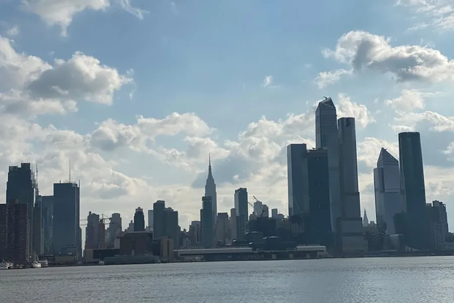 The image shows a daytime silhouette of a city skyline across the water, marked by towering skyscrapers and a scattering of fluffy clouds in the sky.