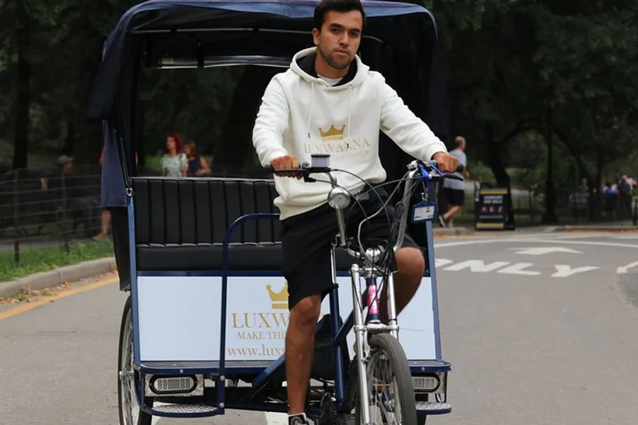 A man is pedaling a pedal-powered rickshaw on a park road, appearing focused on his path ahead.