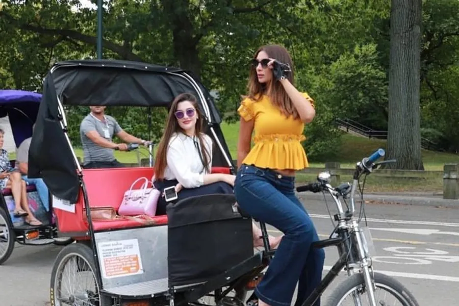Two women, one seated in a pedicab and the other standing beside it, are both wearing sunglasses and appear to be posing for a photo in a park setting.