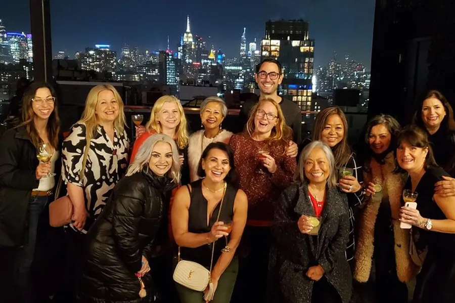 A group of people are smiling and posing for a photo at a night-time rooftop gathering with a city skyline illuminated in the background.
