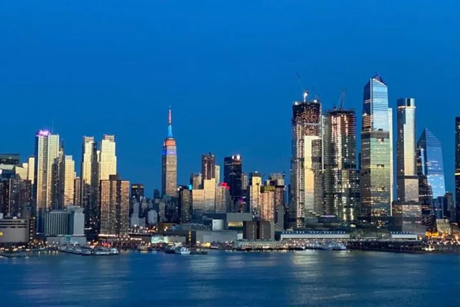 The image shows a panoramic evening skyline of a modern city with illuminated skyscrapers reflecting in the water.