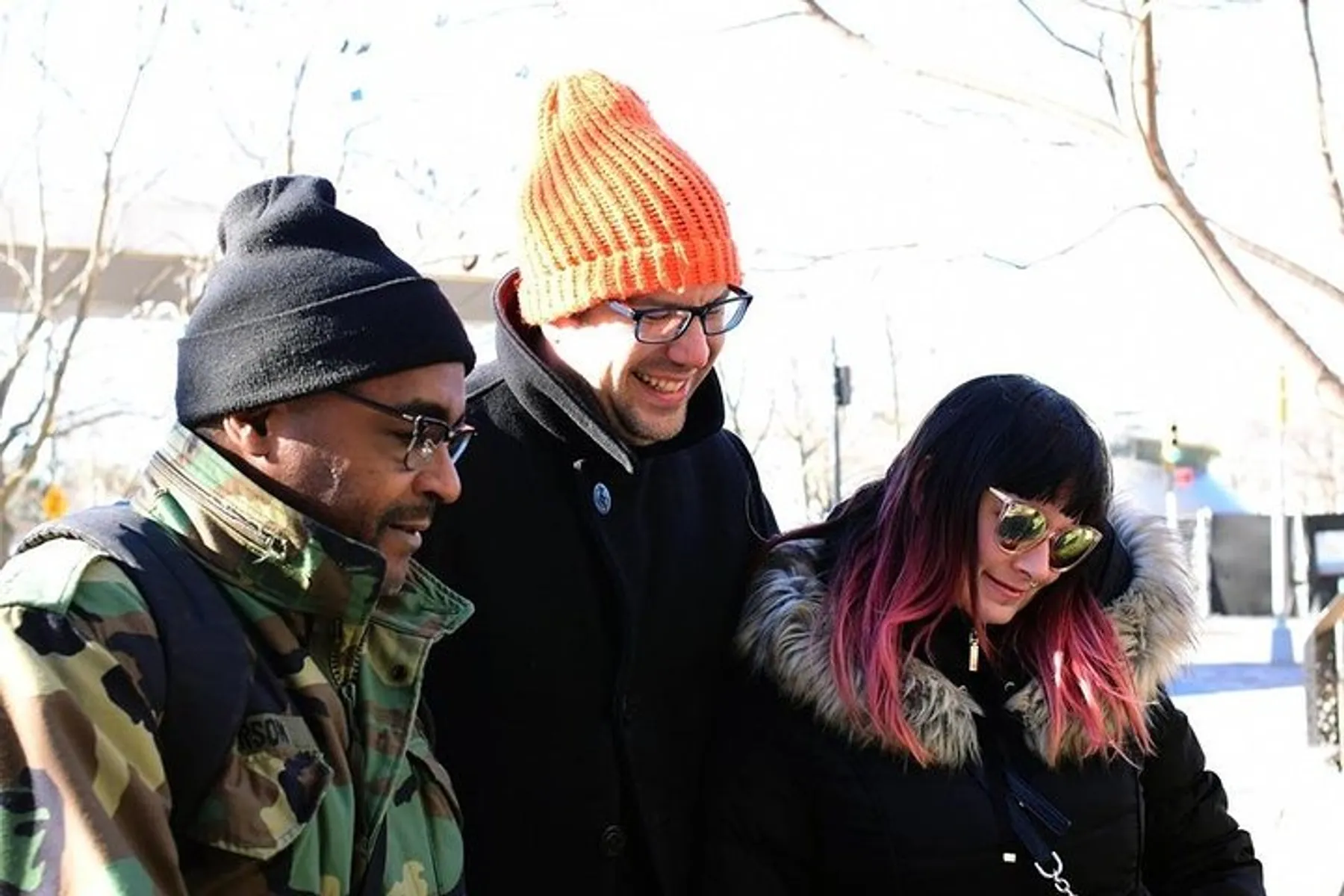 Three people are smiling and looking downward, possibly at something interesting out of frame, while dressed in warm clothes suggesting a cool outdoor setting.