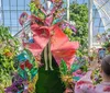 A person is photographing a vibrant botanical display featuring a mannequin dressed in large flower-like attire inside a sunlit greenhouse