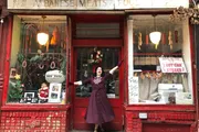 A person in a clown-like outfit is cheerfully posing with arms outstretched in front of an old-fashioned red storefront adorned with holiday decorations and a sign for 