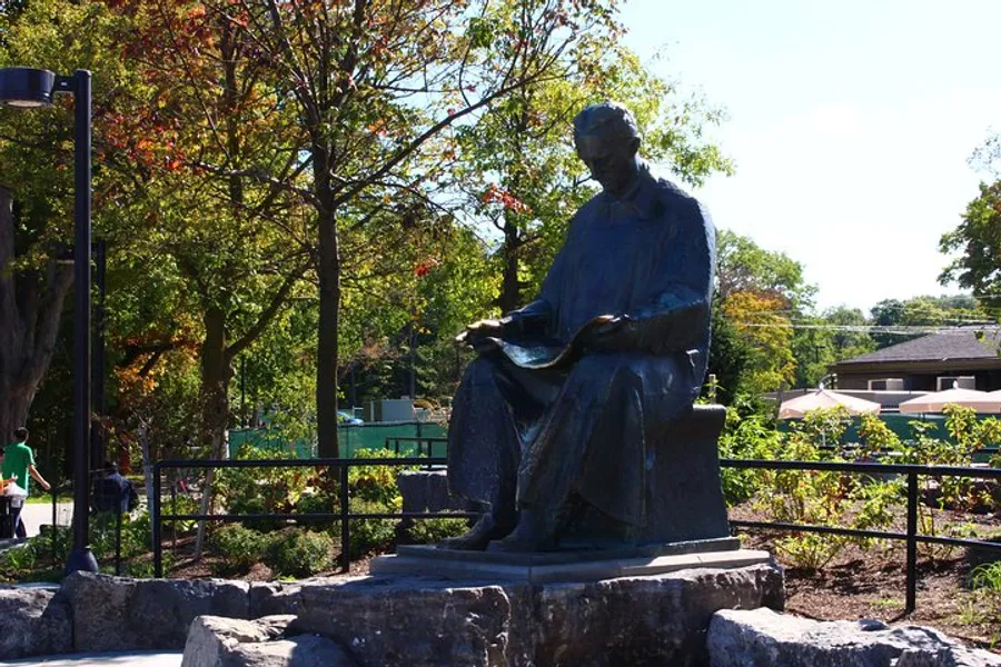 The image shows a bronze statue of a seated figure reading a book, situated in a park-like setting with trees and a clear sky in the background.