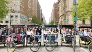 A group of people ride in a line of pedicabs down an urban street flanked by tall buildings.