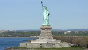 The image shows the Statue of Liberty standing tall on Liberty Island with a clear blue sky in the background and visitors gathered around its base.