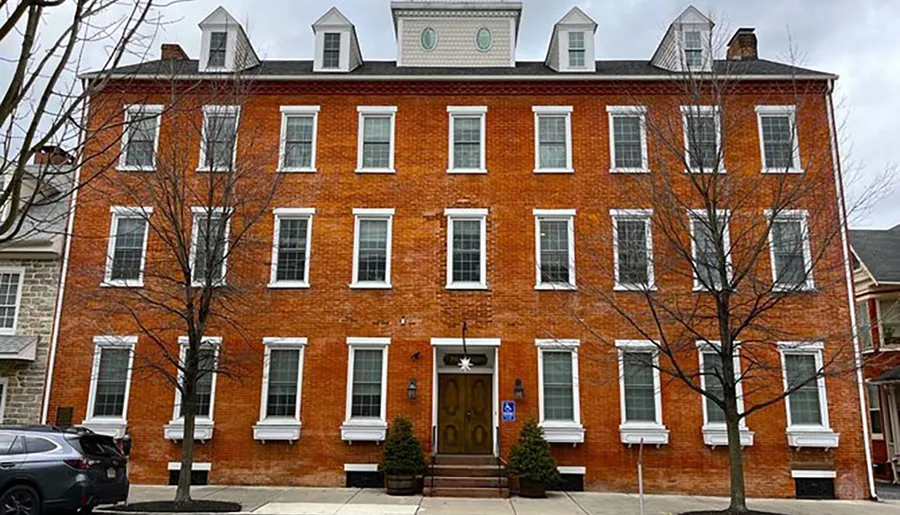 The image shows a symmetrical three-story red brick building with white-trimmed windows and a central entrance flanked by small trees.