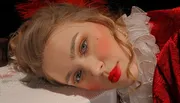 A person with glamorous makeup is lying down, dressed in a red outfit with a ruffled white collar, giving a contemplative look to the camera.