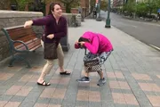 Two women appear to be engaging in playful or mock conflict on a city sidewalk, with one pretending to punch while the other dodges playfully.
