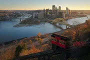 A red funicular car descends along a track overlooking a panoramic view of a city with skyscrapers, bridges, and rivers during sunset.